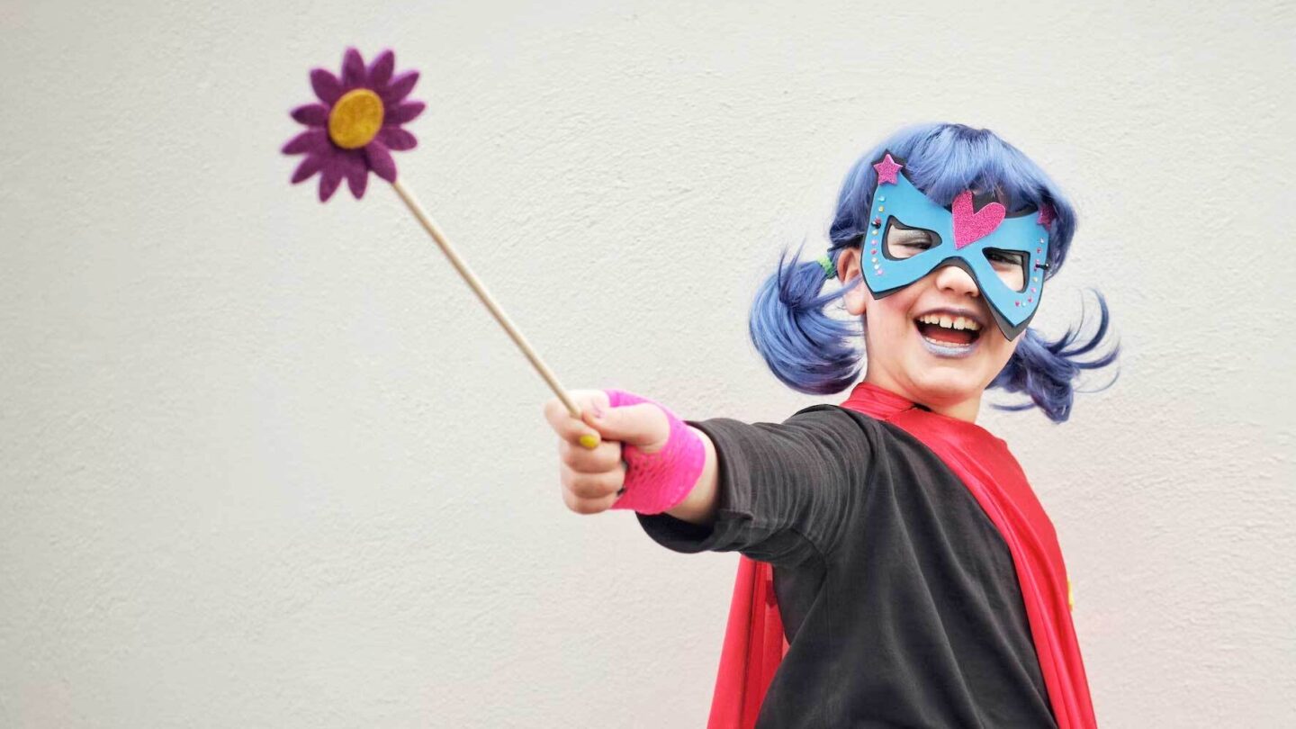A smiling young girl wearing a red cape and a mask over her eyes and holding out a wand with a flower shape at the end.