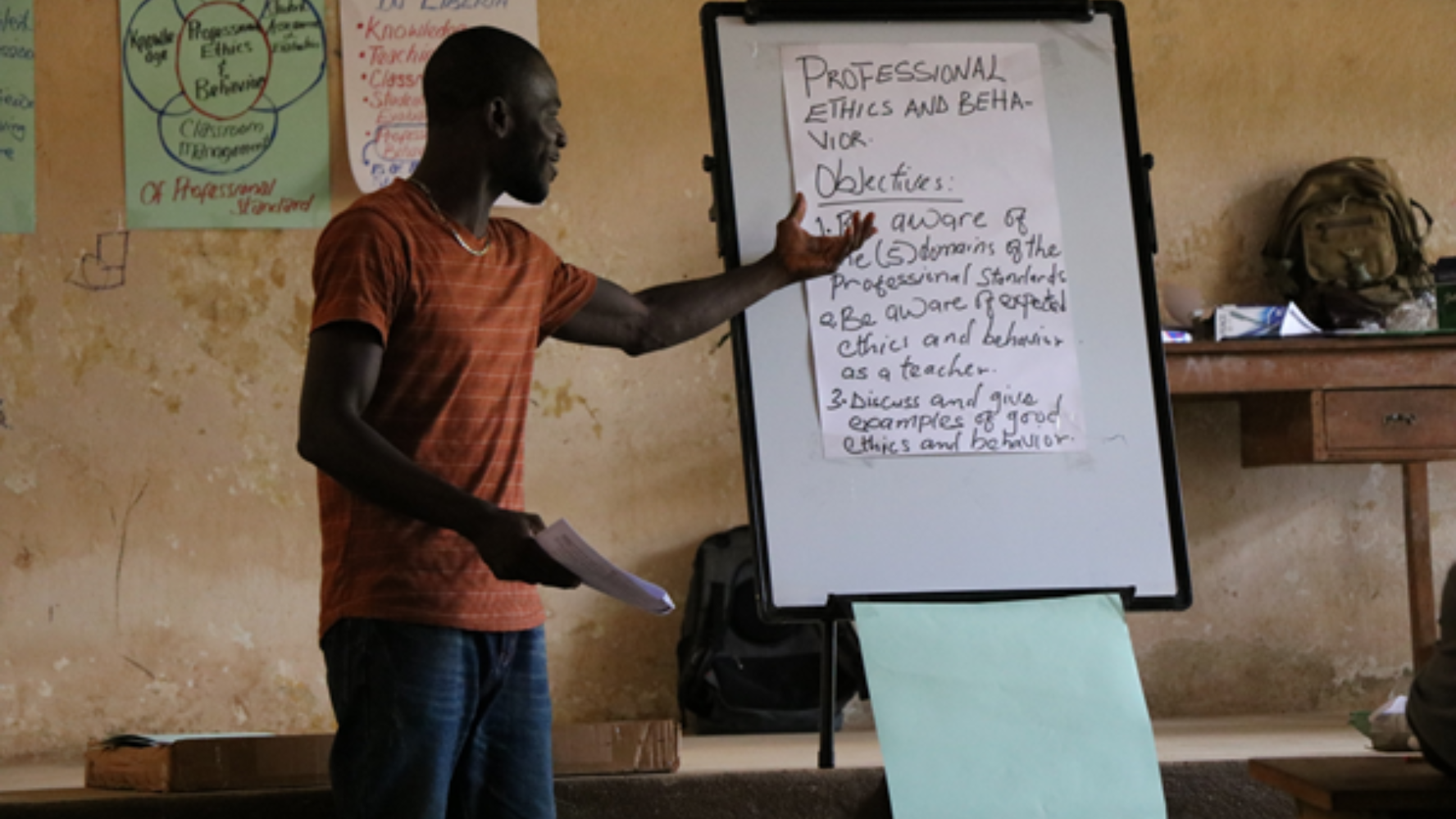 A man in a rural schoolroom standing in front of a whiteboard, on which is written 'Professional ethics and behaviour'.