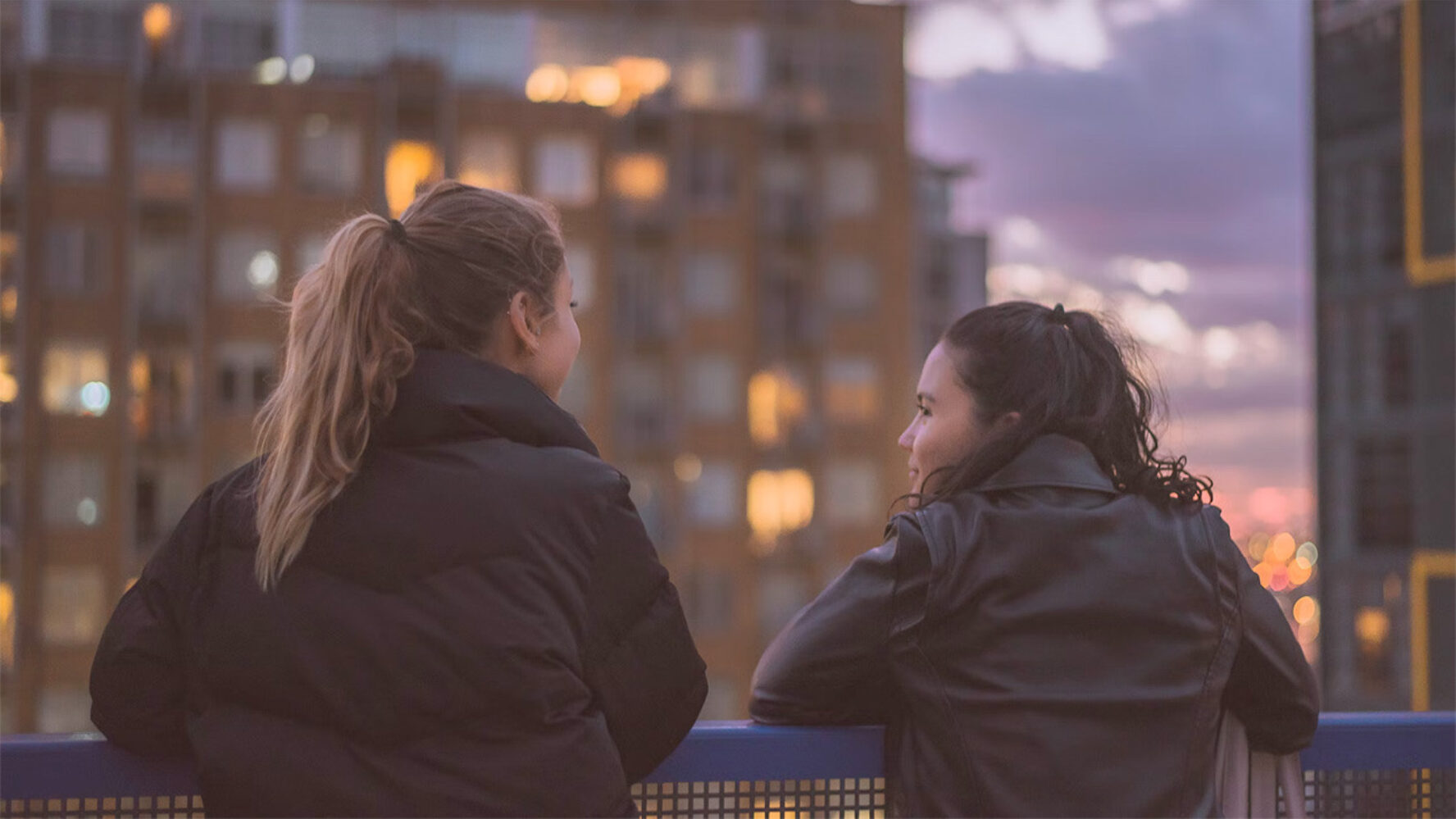 Two young women facing away and looking out over a city at dusk