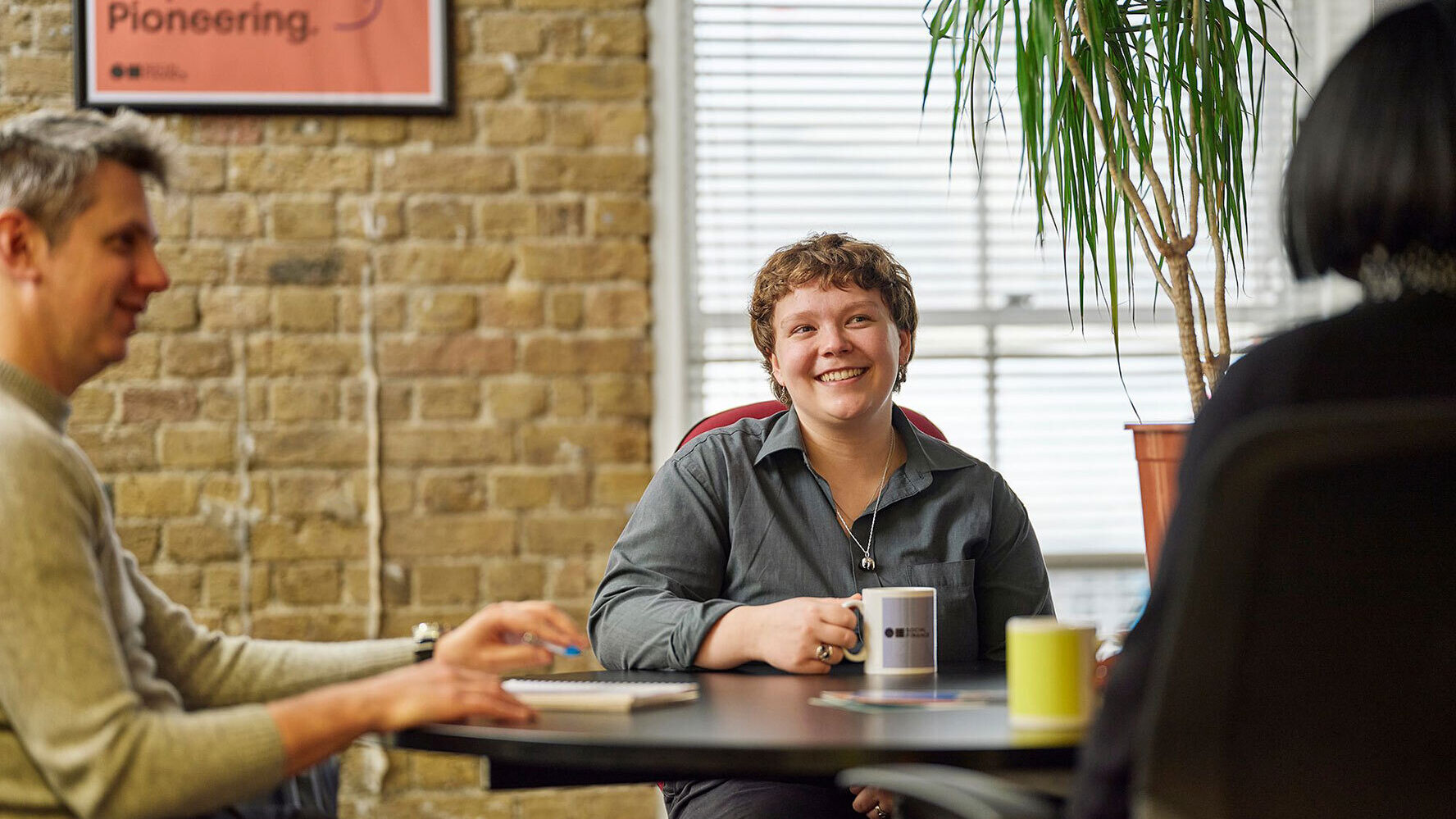 Three people sat round a table in an office, sharing a coffee and talking.